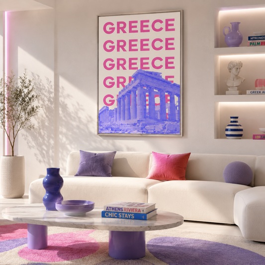 A modern living room featuring a large vertical wall art print with the word 'GREECE' repeated in pink above a blue graphic of the Parthenon. The room has a white sectional sofa with pink and purple pillows, a pink armchair, and a round pink and purple rug.