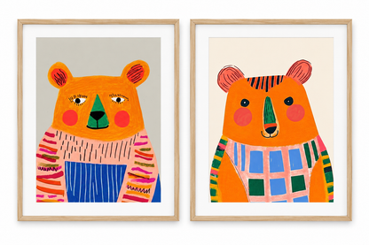 Two framed whimsical bear art prints side by side, featuring orange bears in colorful striped and plaid sweaters, set in light wood frames against a white background.