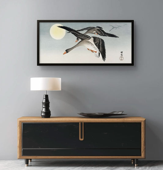 Framed artwork of two geese flying over a moonlit sky above a wooden cabinet with a lamp.