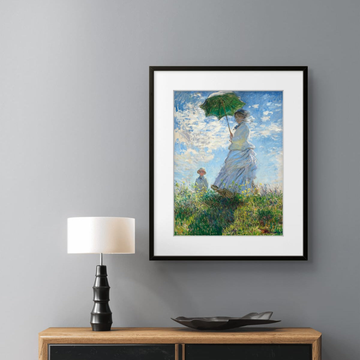 A framed art print of Claude Monet's 'Woman with an Umbrella' hanging on a gray wall above a wooden and black sideboard with a lamp.